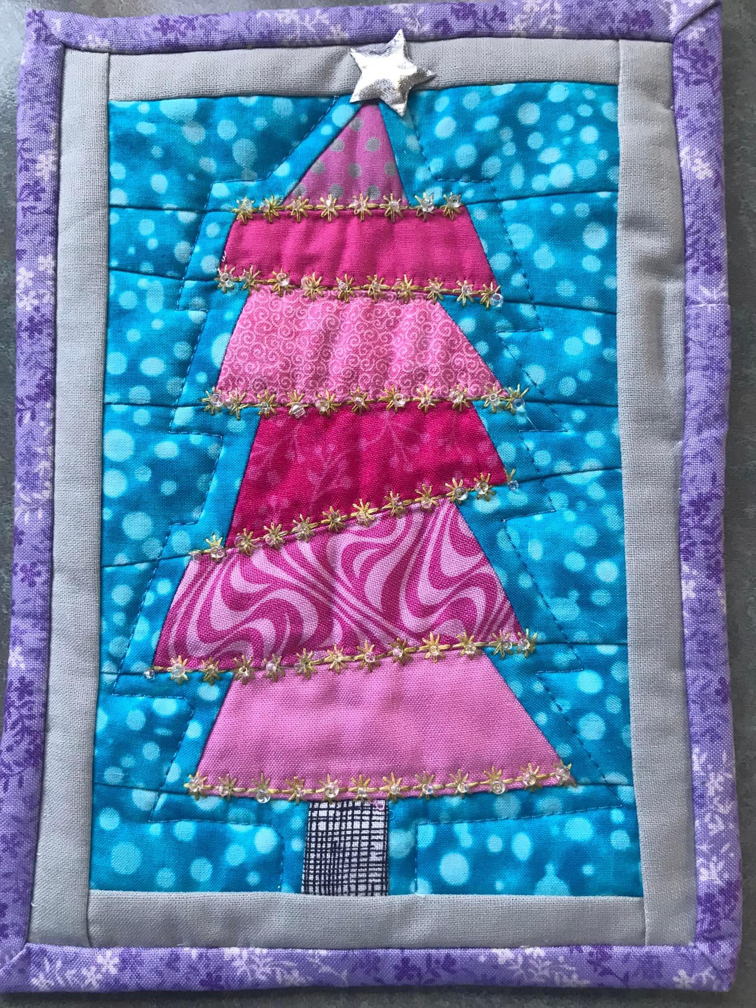 A small, square quilt with a purple border. The quilt features a pink Christmas tree with a silver star on top. The tree is decorated with gold thread and is set against a blue background with white polka dots.