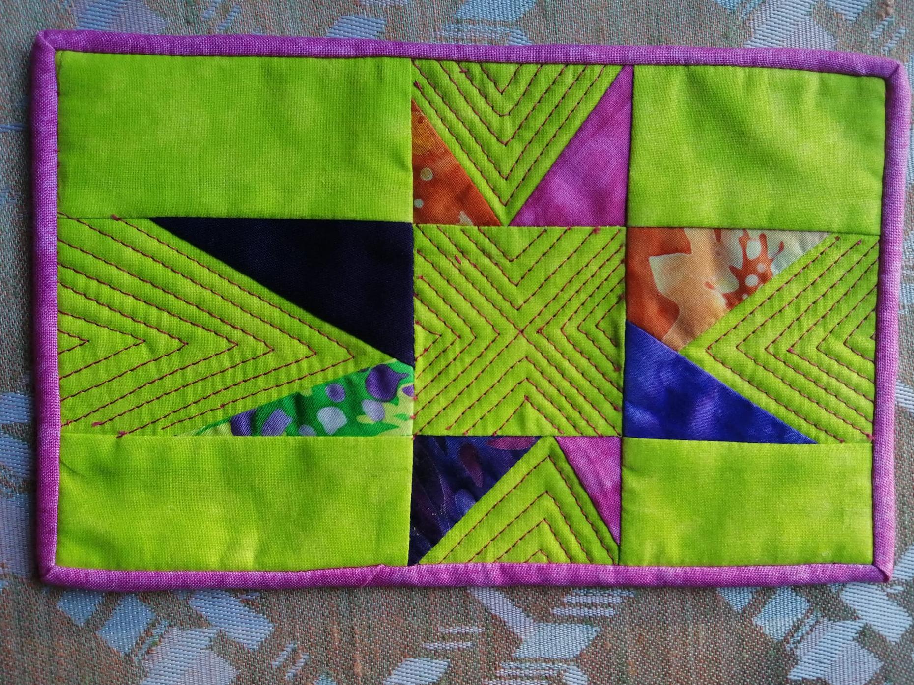 A small, rectangular quilt with a green background and a pink border. The quilt features a colorful patchwork design with triangles in various colors like pink, orange, blue, and black. The triangles are stitched with a decorative zigzag pattern.