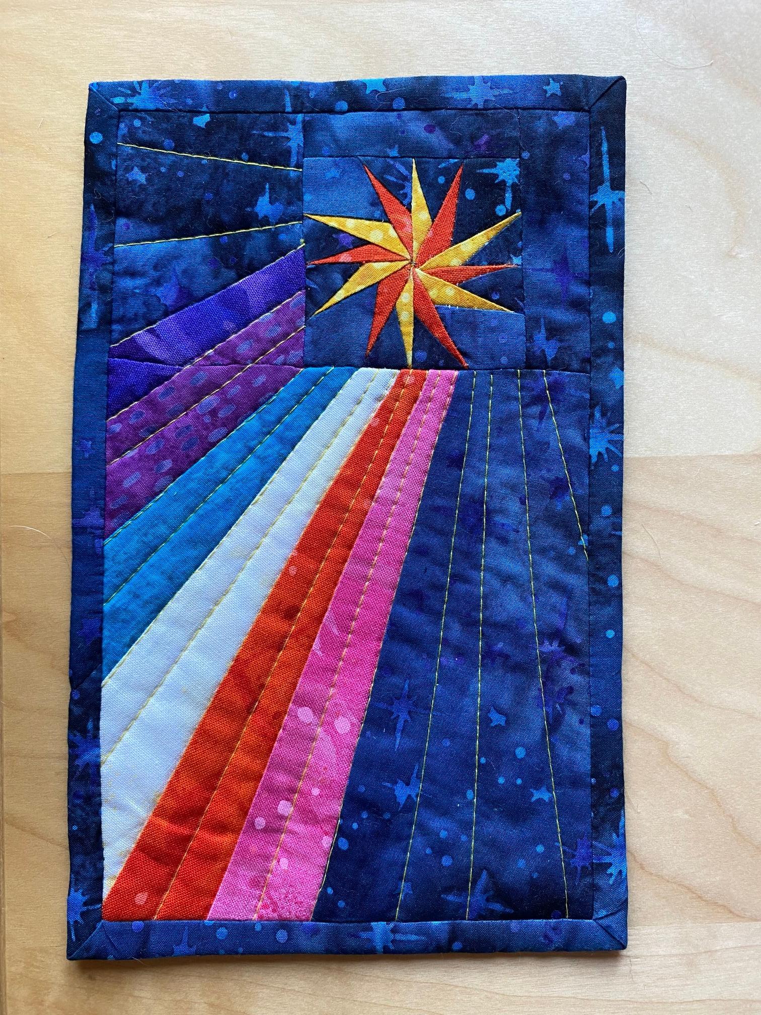 A square quilt with a dark blue background. The quilt features a bright, multi-colored star in the top center, with rays of light extending downwards in various colors, including purple, blue, white, red, and pink. The background is quilted with a subtle, linear pattern.