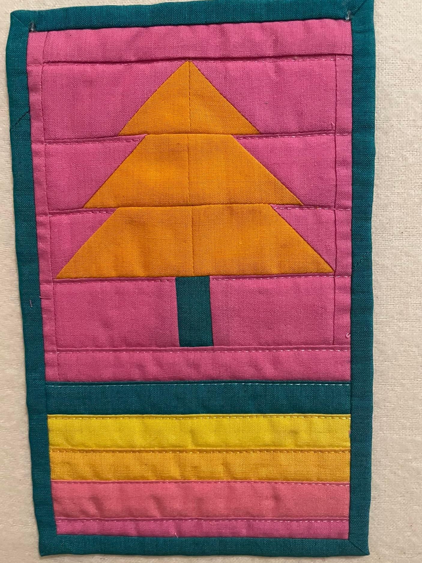 A small, rectangular quilt with a green border. The quilt features a stylized Christmas tree made of orange triangles on a pink background. The base of the quilt is a gradient of pink, orange, and yellow.