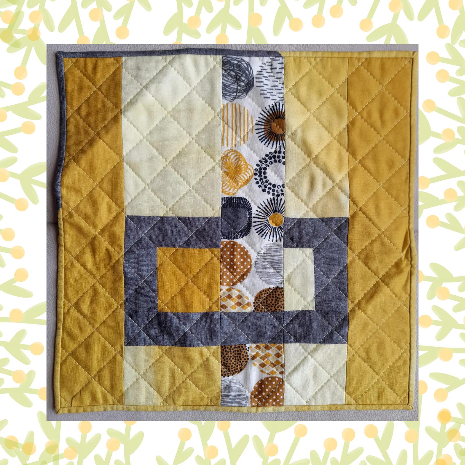 A square quilt with a patchwork design in shades of yellow and grey. The quilt features a central panel with a geometric pattern of squares and rectangles, surrounded by a border of solid yellow fabric. The background is quilted with a diamond pattern.