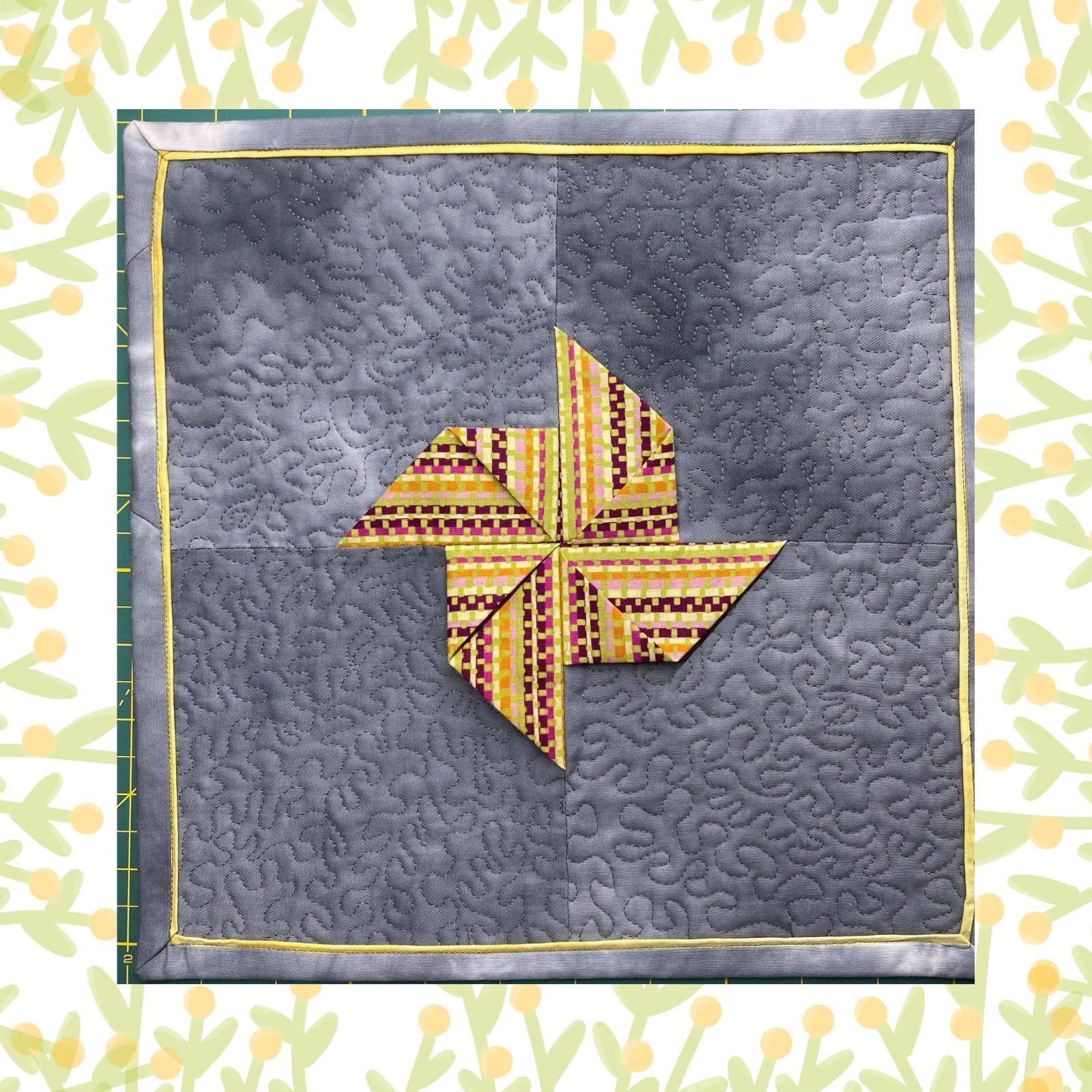 A square quilt with a grey background and a yellow border. The quilt features a colourful pinwheel design in the centre, made up of triangles in various shades of pink, yellow, and orange. The background is quilted with a stipple pattern.