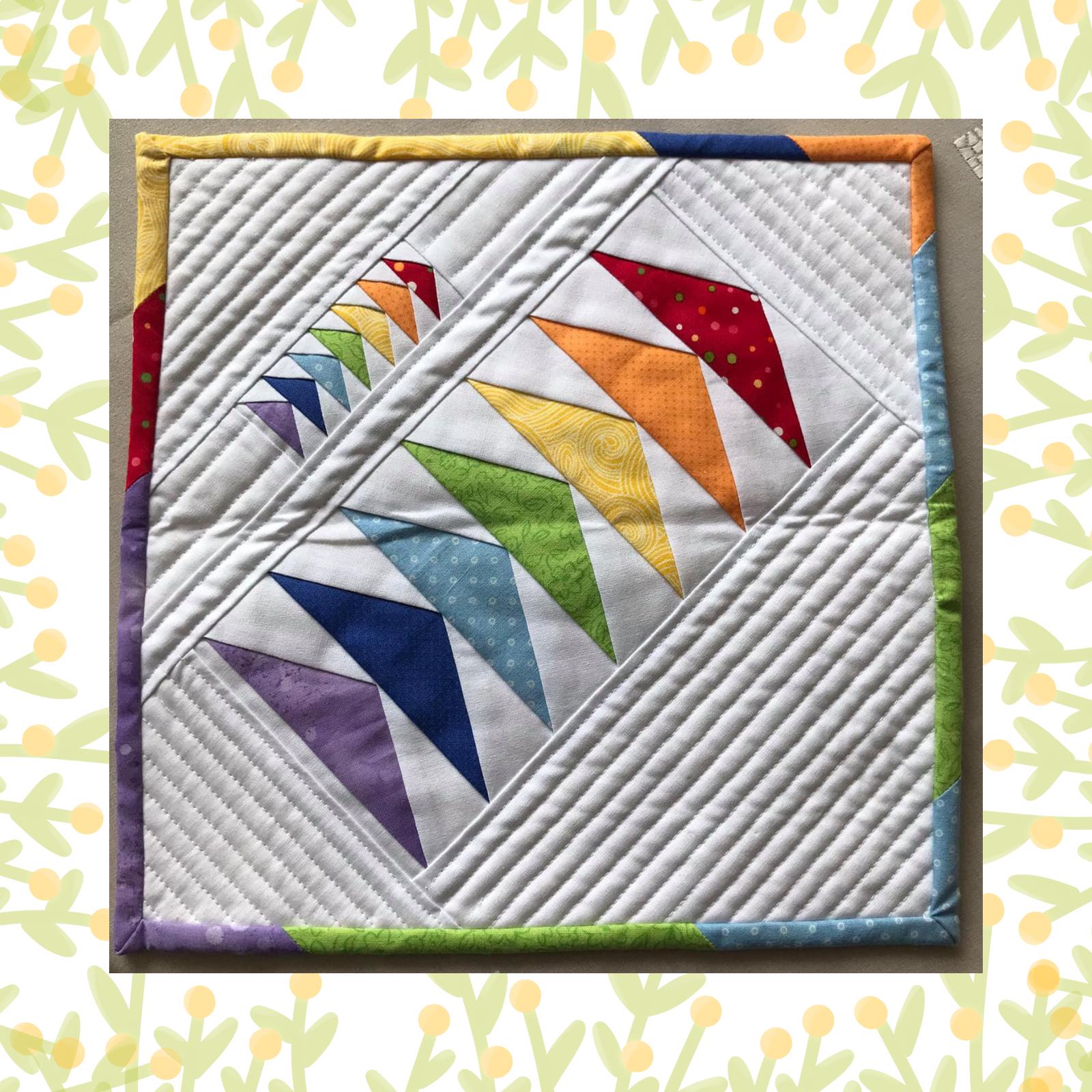 A square quilt with a white background and a colourful border. The quilt features a rainbow design made up of triangles in various colours, including red, orange, yellow, green, blue, and purple. The background is quilted with diagonal lines.