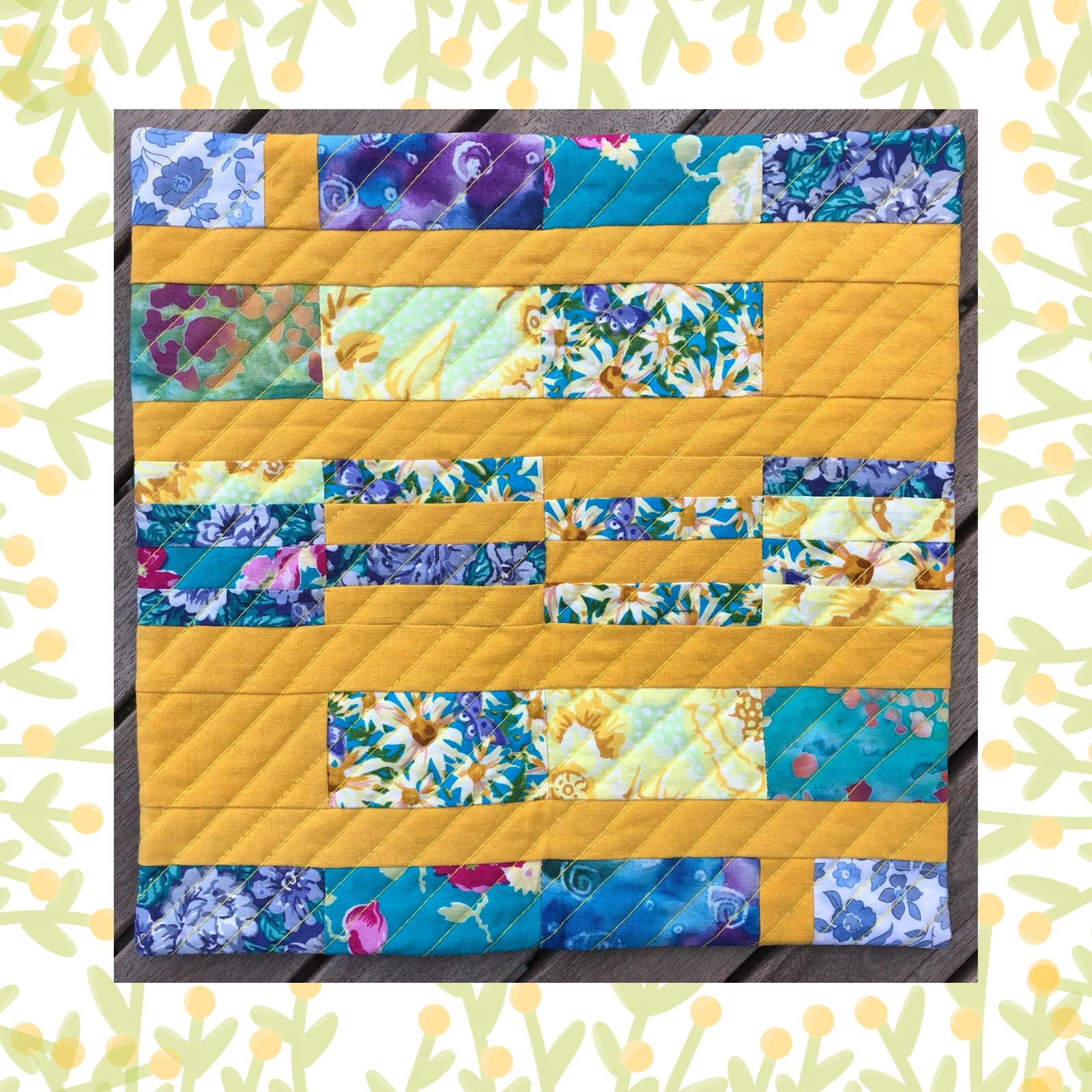 A square quilt with a yellow background. The quilt features a patchwork design with horizontal strips of fabric in various colours and patterns, including floral prints and solid colours. The background is quilted with a diagonal line pattern.