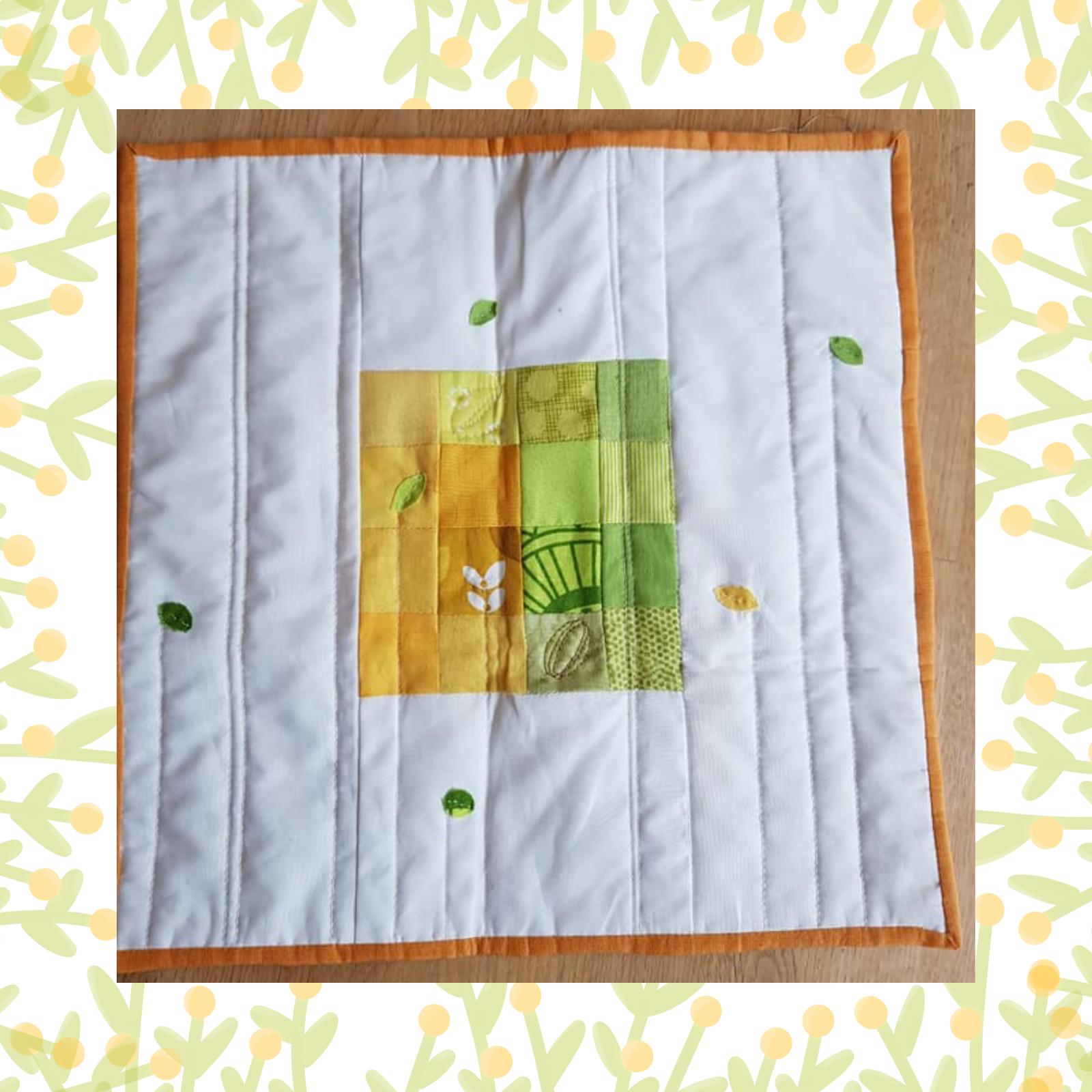A square quilt with a white background and an orange border. The quilt features a patchwork design in shades of green and yellow, with small, embroidered flowers scattered around. The background is quilted with vertical lines.