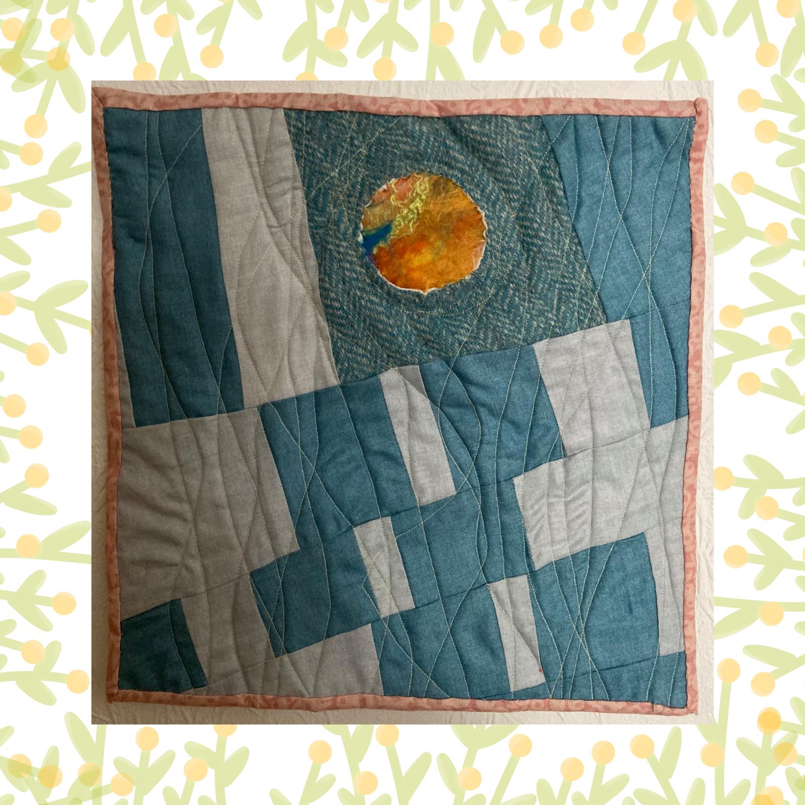 A square quilt with a predominantly blue and grey colour scheme. The quilt features a patchwork design with geometric shapes, including squares and rectangles. A circular, textured orange patch is placed in the centre of the quilt. The background is quilted with wavy lines. The quilt is framed by a pink border.