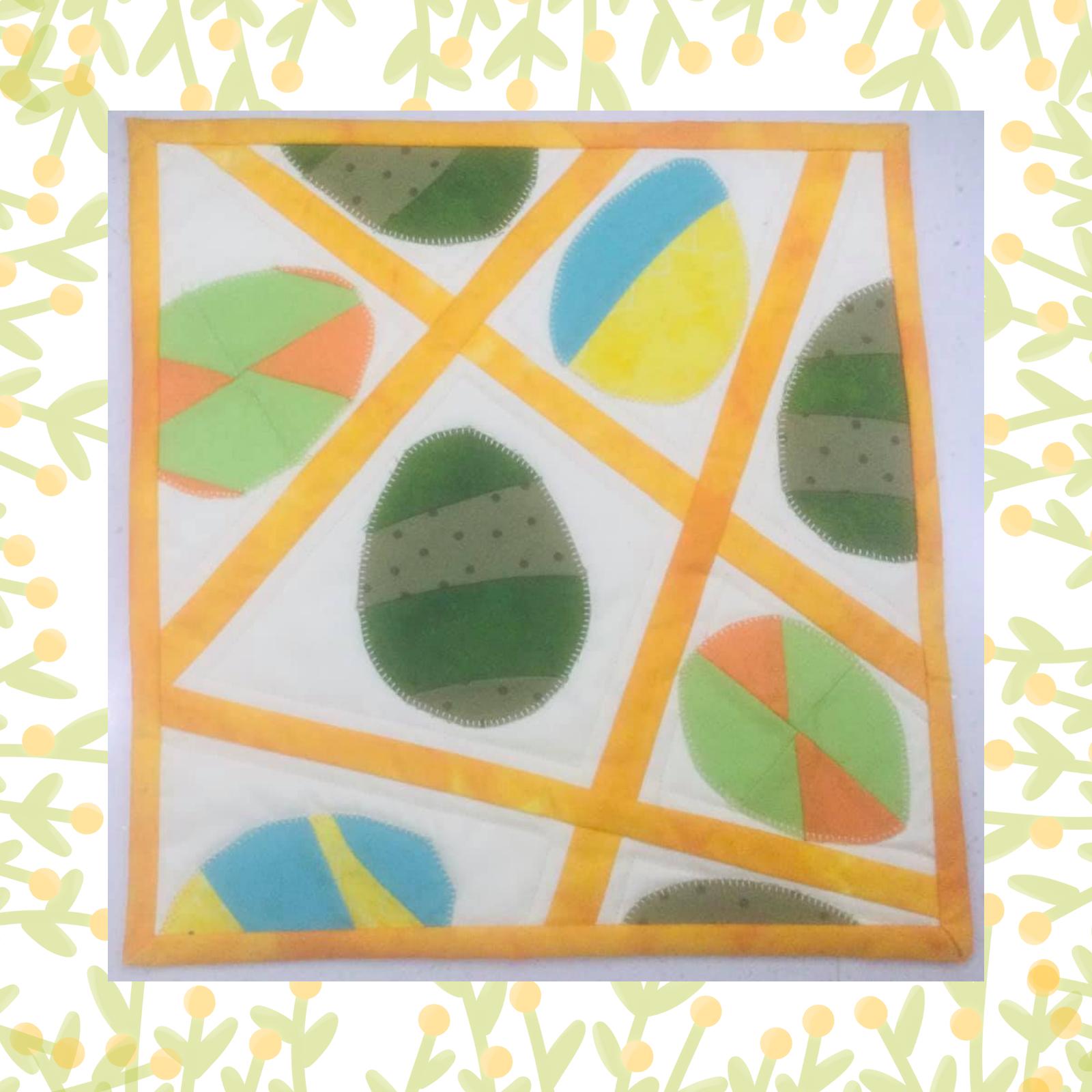 A square quilt with a white background and a yellow border. The quilt features colourful eggs scattered across the background, with yellow strips of fabric intersecting them. The eggs are in various colours and patterns, including solid colours, polka dots, and stripes.