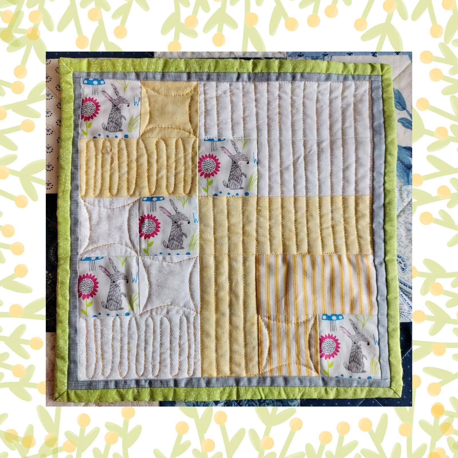 A square quilt with a yellow background and a green border. The quilt features a patchwork design with squares and rectangles in various shades of yellow and grey. There are small, embroidered images of rabbits and mushrooms throughout the quilt. The background is quilted with a linear pattern.