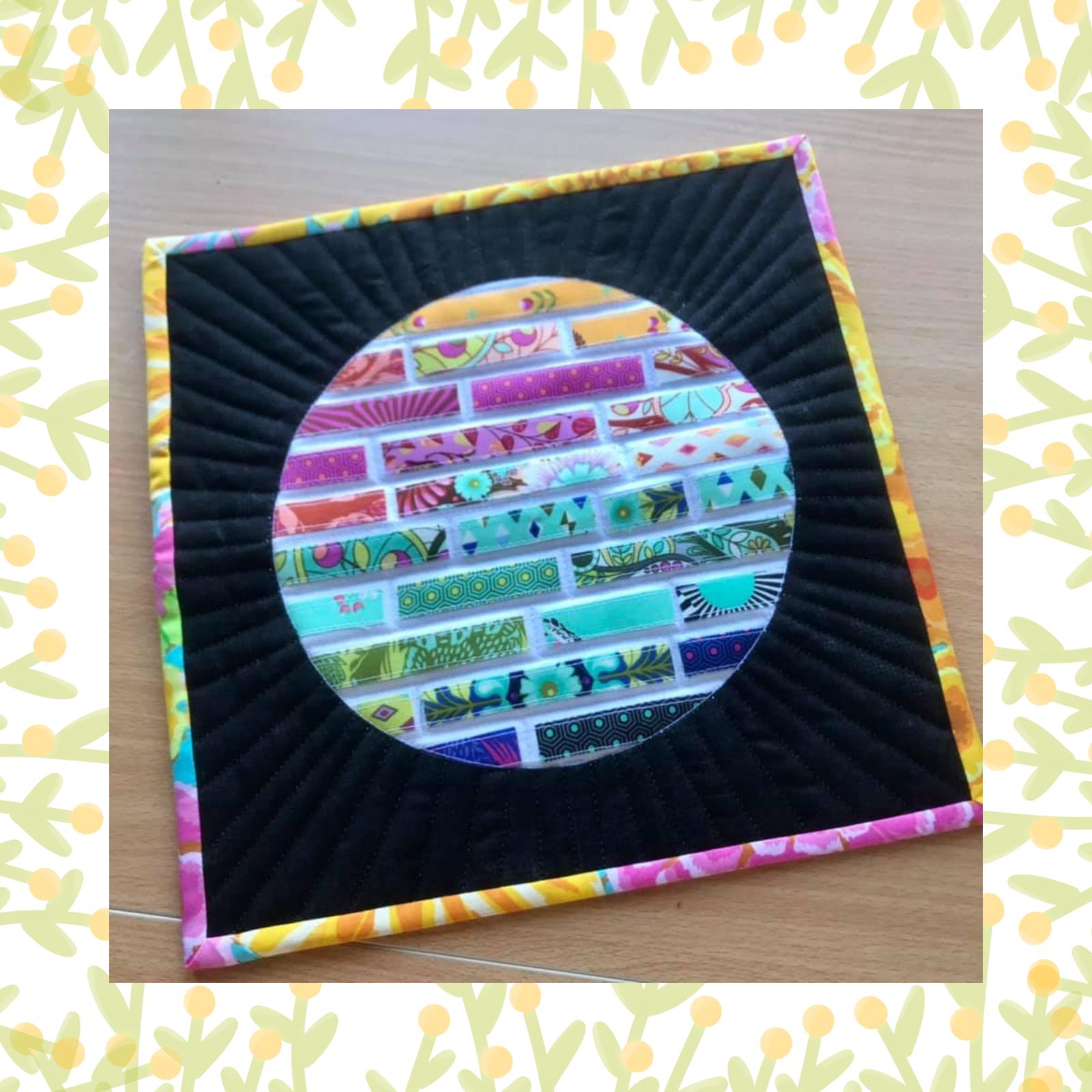 A square quilt with a black background and a colourful border. The centre of the quilt features a circular design made up of small, rectangular pieces of fabric in various colours and patterns. The background is quilted with radiating lines.