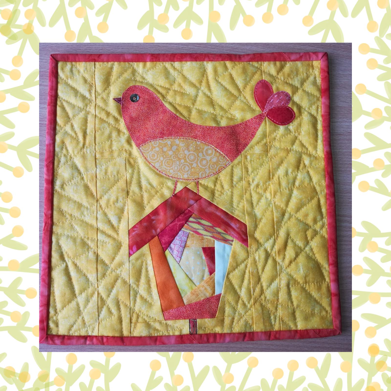 A square quilt with a yellow background and a red border. The quilt features a red bird perched on a colourful birdhouse. The birdhouse is made up of various shapes and colours, including orange, yellow, and red. The background is quilted with a triangular pattern