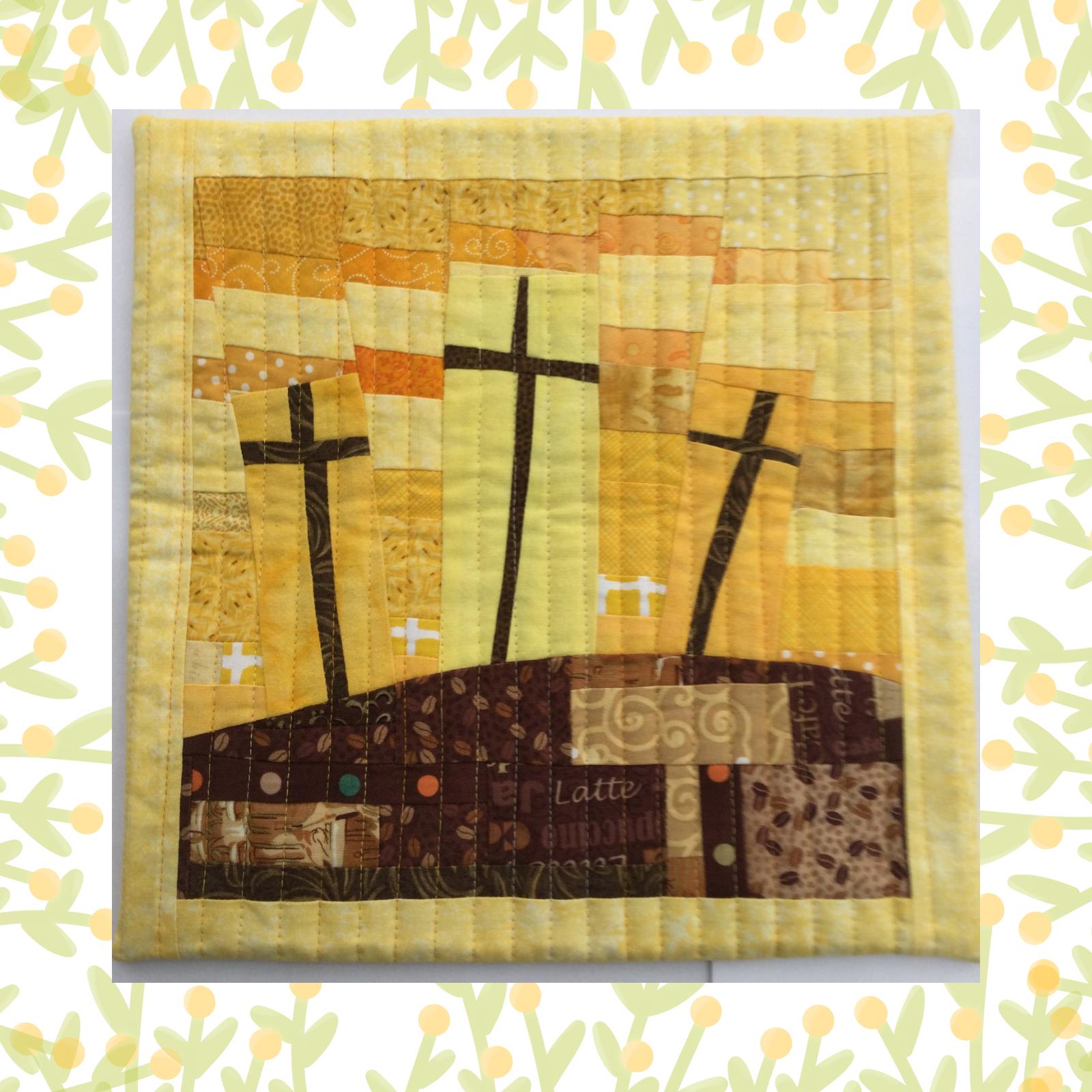A square quilt with a yellow background and a brown border. The quilt features three crosses on a hill, with a sunset-like sky behind them. The ground is depicted with a patchwork of brown and orange fabrics. The word "Latte" is visible in a small, colourful patch of fabric.