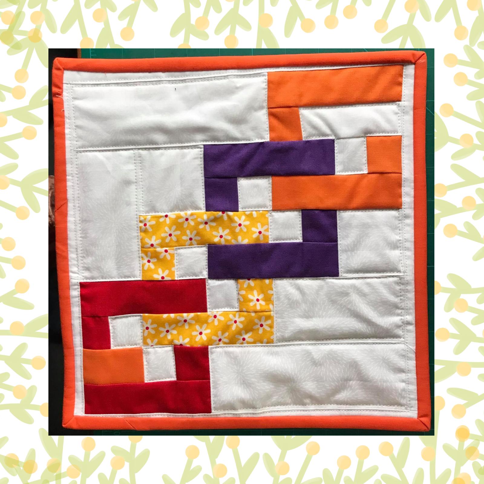 A square quilt with a white background and an orange border. The quilt features a colourful, geometric design with overlapping shapes in red, orange, purple, and yellow. Some of the shapes have a floral pattern. The background is quilted echoing the pieces of the quilt..