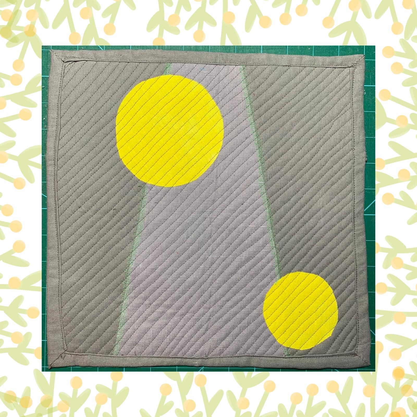 A square quilt with a grey background and a green border. The quilt features two large, yellow circles, one larger than the other, positioned diagonally on the grey background. The background is quilted with a linear pattern.