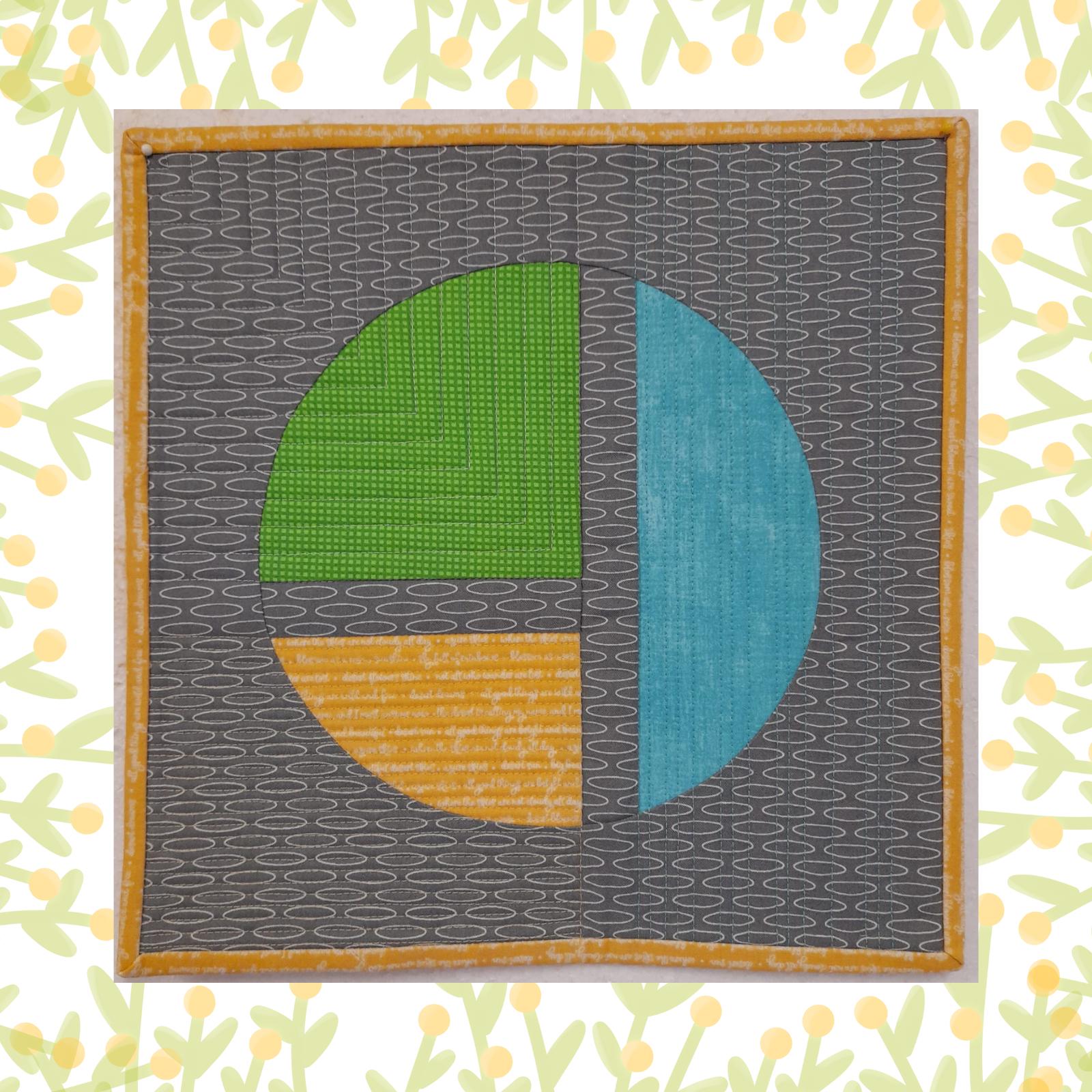 A square quilt with a grey background and a yellow border. The quilt features a circle divided into four sections, each with a different colour and texture: green, yellow, blue, and grey. The background is quilted with straight lines.