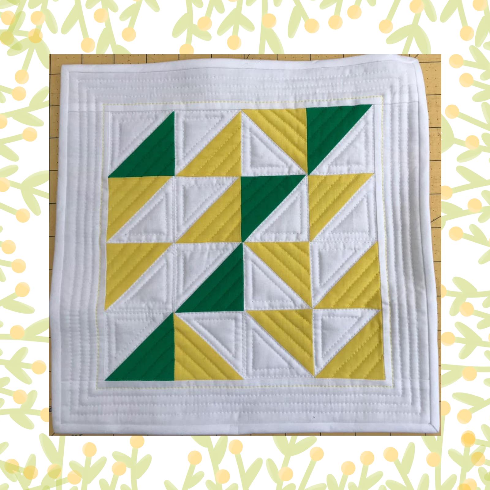 A square quilt with a white background. The quilt features a geometric pattern of alternating green and yellow triangles, creating a visually interesting design. The background is quilted with a linear pattern. The quilt is framed by a white border.