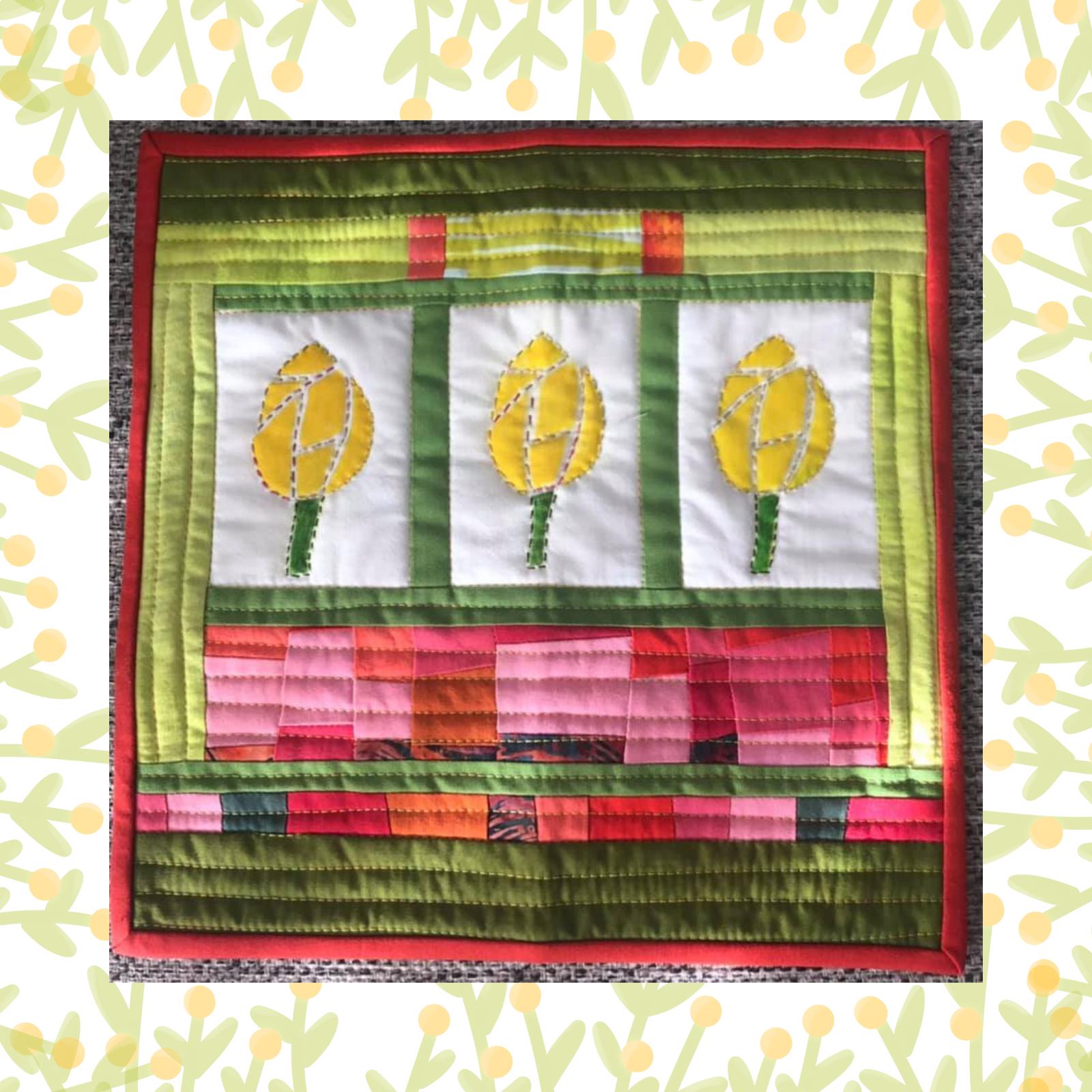 A square quilt with a green background and a red border. The quilt features three yellow tulips embroidered on a white background. Below the tulips, there is a colourful patchwork design in shades of pink and orange. The background is quilted with a linear pattern.
