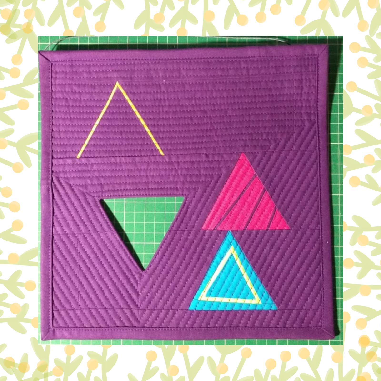 A square quilt with a purple background and a green border. The quilt features three triangles of different colours: yellow, pink, and blue. The triangles overlap each other, creating a dynamic composition. The background is quilted with a linear pattern.