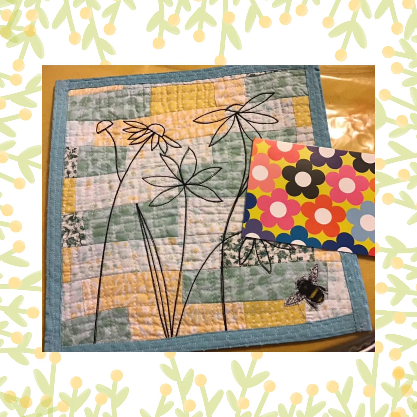 A square quilt with a patchwork design in blue, yellow, and white. The quilt features embroidered flowers and a small, embroidered bee. Next to the quilt is a card with a colourful, geometric pattern. The quilt is framed by a green border with yellow flowers.