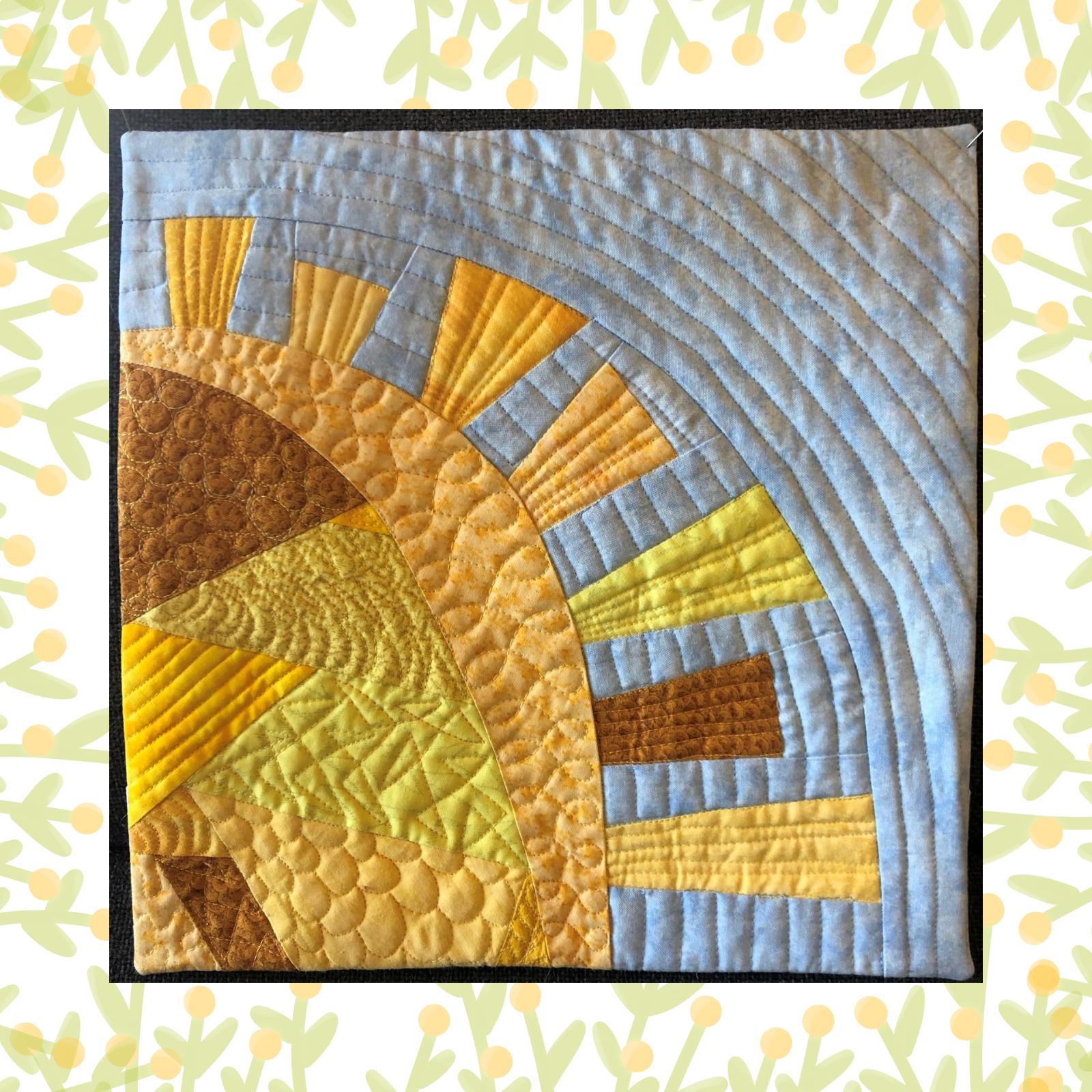 A square quilt with a blue background. The quilt features a large, yellow sun with orange and brown rays extending outwards. The sun and rays are made of fabric pieces sewn together, with quilted details adding texture. The background is quilted with curved lines.