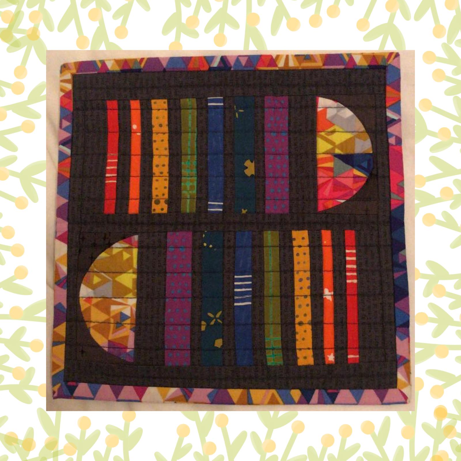 A square quilt with a dark grey background. The quilt features a colourful geometric design with vertical and curved shapes in various colours, including red, orange, yellow, green, and blue. The shapes are arranged in a symmetrical pattern. The quilt is framed by a grey border.