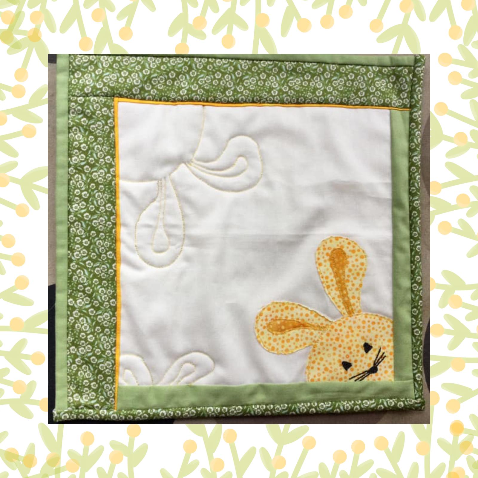 A square quilt with a white background and a green border. The quilt features a cute, embroidered bunny peeking out from the corner.