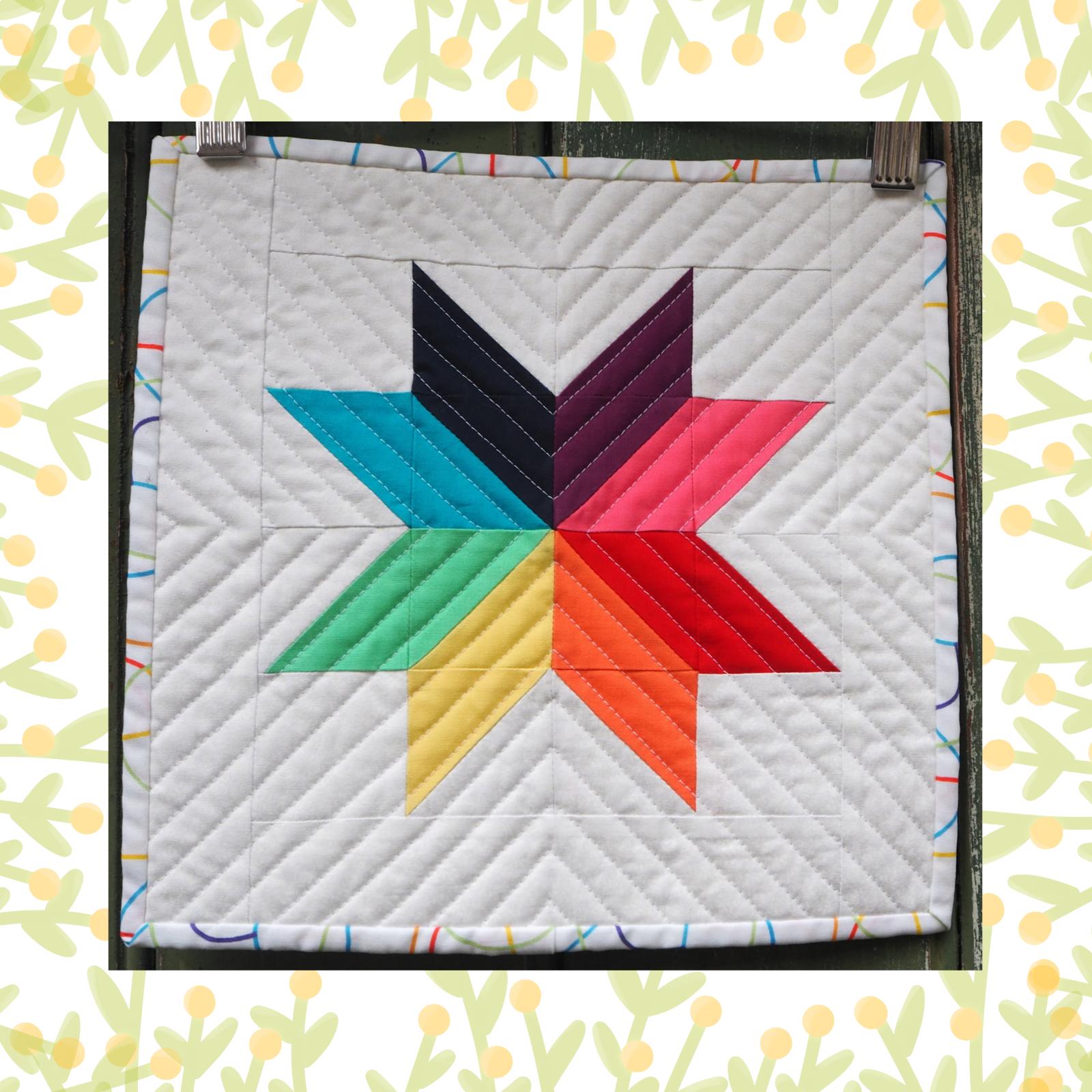 A square quilt with a white background. The quilt features a colourful, eight-pointed star made up of triangles in various colours, including red, orange, yellow, green, blue, purple, and pink. The background is quilted with a chevron pattern. The quilt is framed by a colourful, striped border.