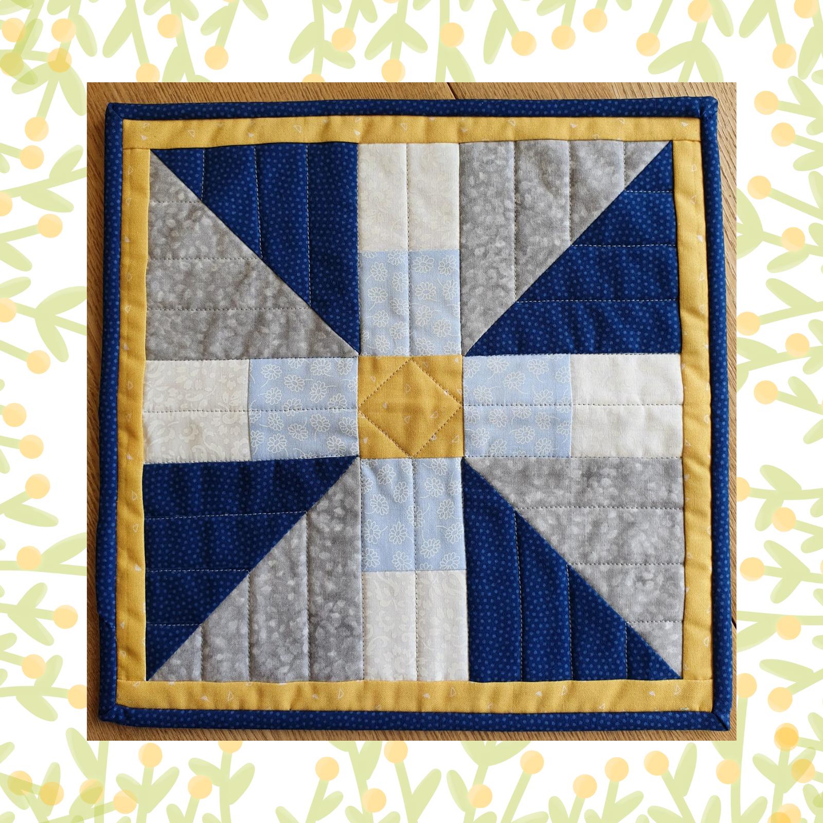 A square quilt with a yellow border. The quilt features a colourful, geometric design with triangles in shades of blue, grey, and white. The centre of the quilt has a small, yellow square. The background is quilted with a linear pattern.
