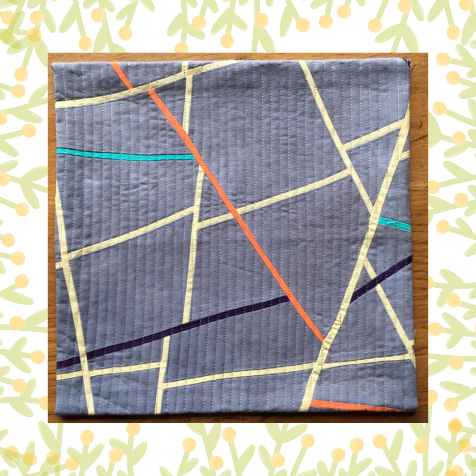 A square quilt with a grey background. The quilt features a geometric design with intersecting lines in various colours, including orange, yellow, purple, and blue. The background is quilted with a linear pattern.