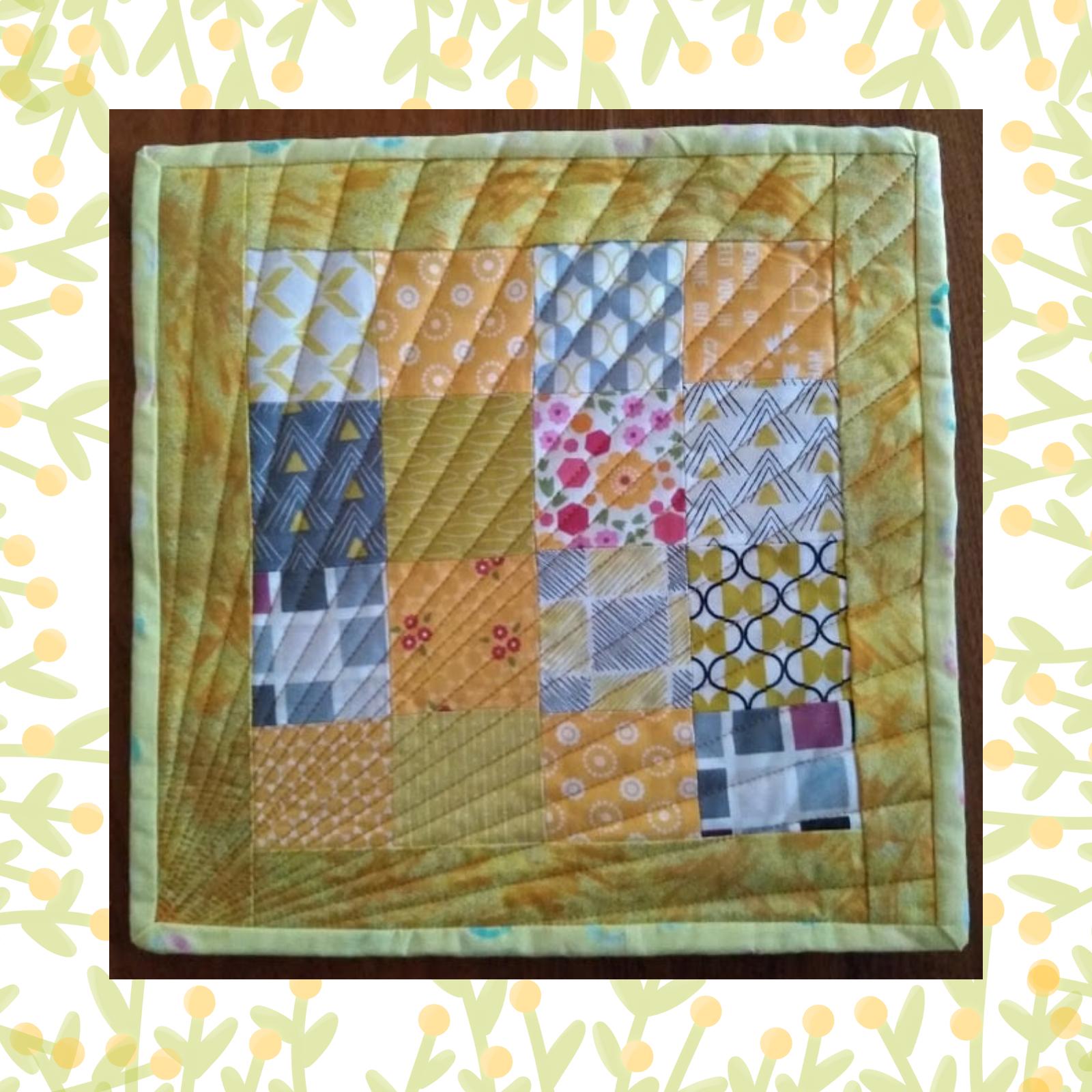 A square quilt with a yellow background and a yellow border. The quilt features a patchwork design with squares in various colours and patterns, including grey, pink, and yellow. The background is quilted with radiating lines.