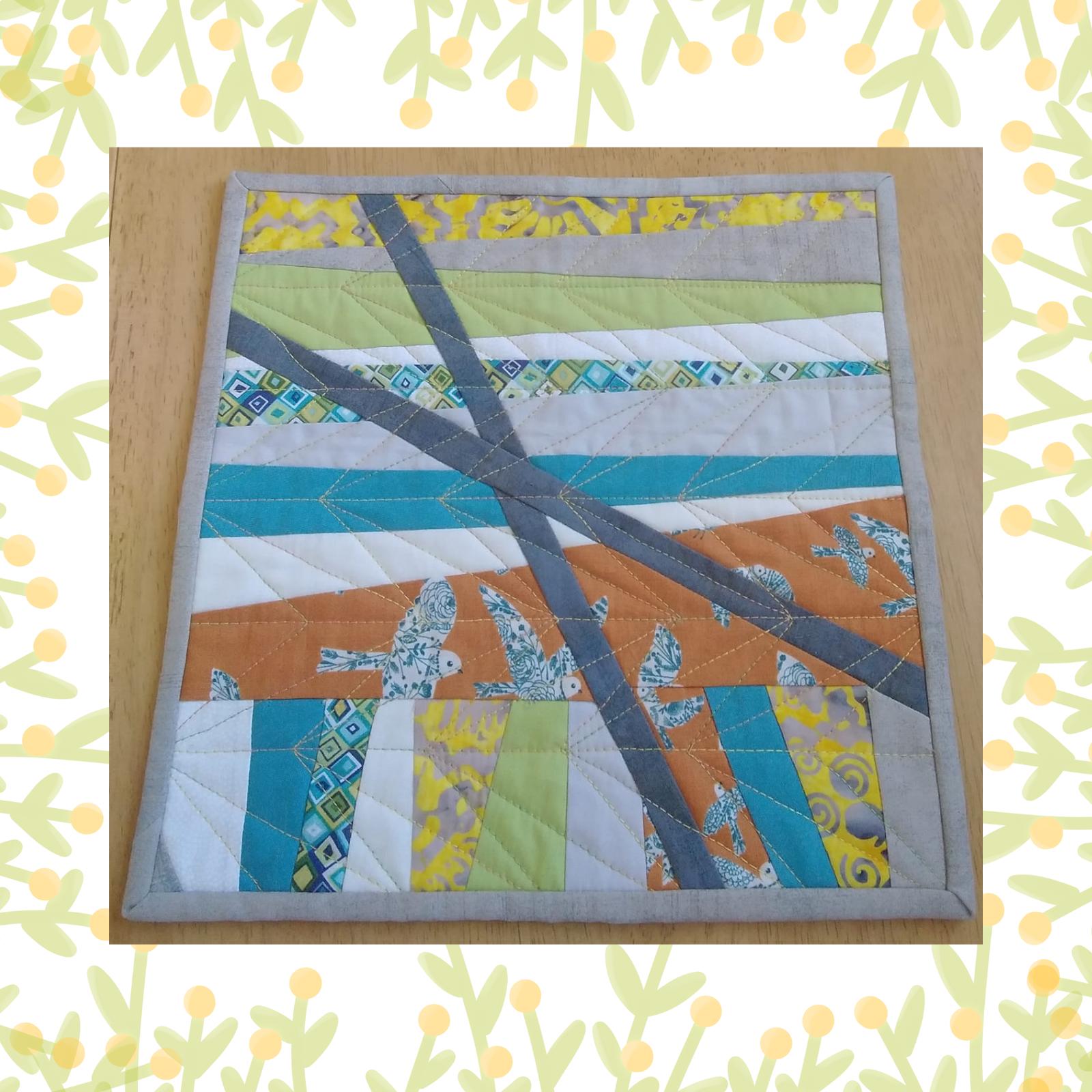 A square quilt with a patchwork design in shades of blue, green, and orange. The quilt features diagonal stripes of fabric, with smaller squares and rectangles creating a geometric pattern. There are also small, grey birds scattered throughout the quilt.