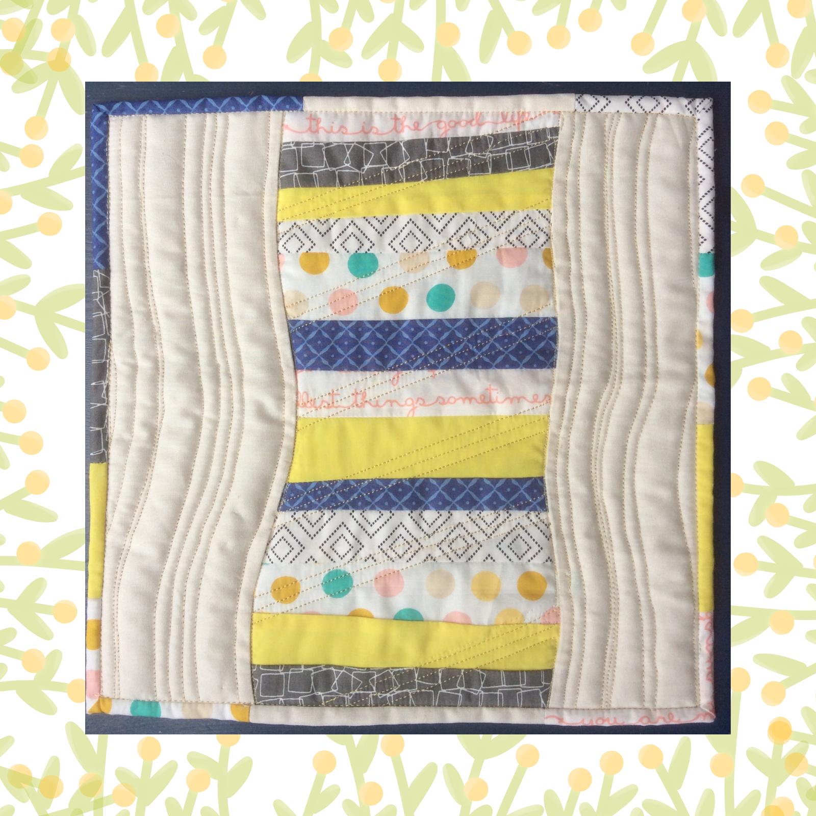 A square quilt with a blue background and a blue border. The quilt features a colourful, abstract design with vertical strips of fabric in various colours and patterns, including yellow, blue, and grey. The background is quilted with a linear pattern.