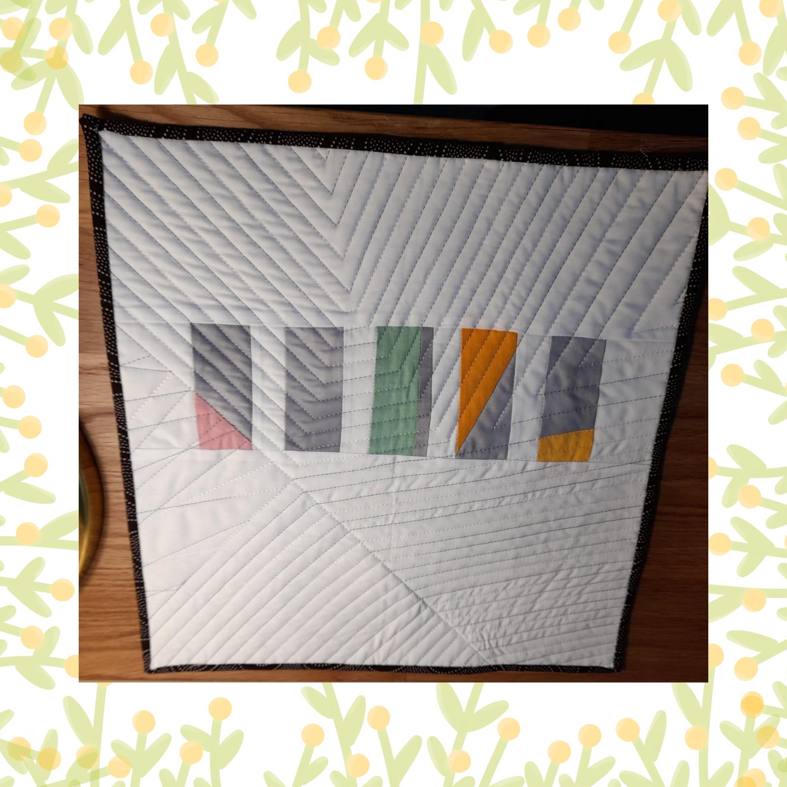 A square quilt with a white background and a black border. The quilt features a row of colourful rectangles in shades of pink, green, yellow, and grey. The background is quilted with a diagonal line pattern.