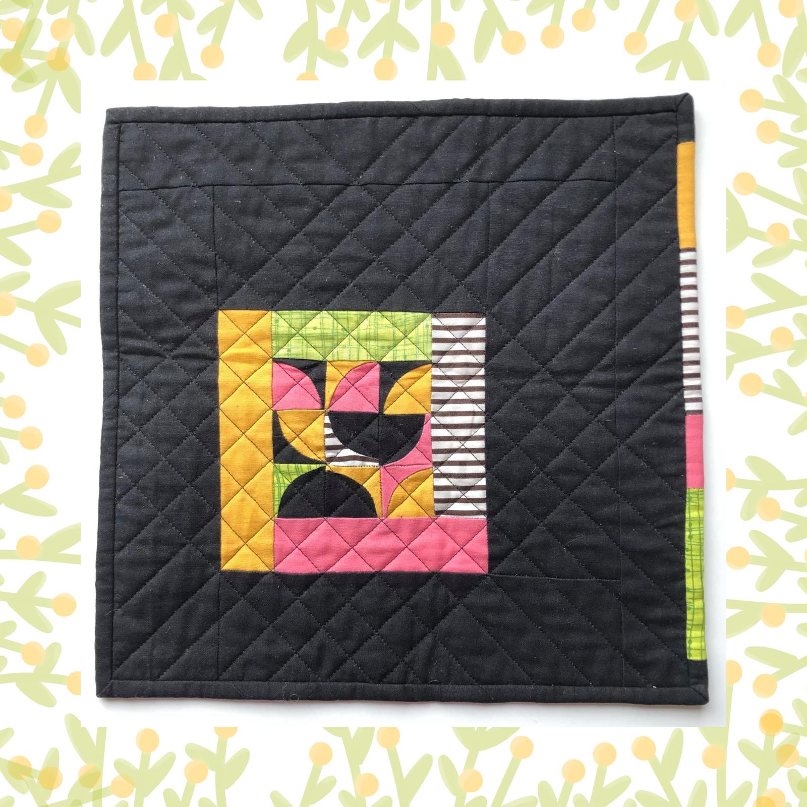 A square quilt with a black background and a yellow border. The quilt features a colourful patchwork design in the centre, with shapes in pink, yellow, and green. The background is quilted with a diamond pattern.
