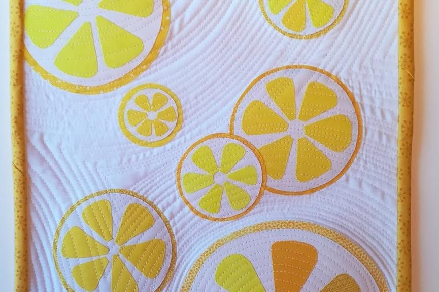 A square quilt with a white background and a yellow border. The quilt features multiple lemon slices in various sizes, all in shades of yellow. The background is quilted with a wavy pattern