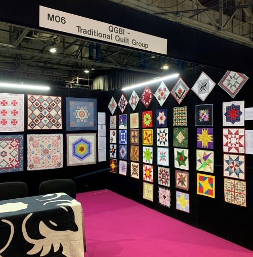 A selection of quilts displayed on an exhibition stand under a sign 'QGBI - Traditional Quilt Group'