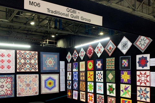 A selection of quilts displayed on an exhibition stand under a sign 'QGBI - Traditional Quilt Group'