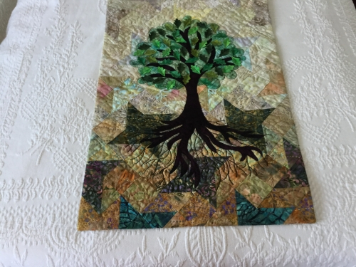 Rectangular quilt with a background of neutral tones and a tree with green leaves in the foreground