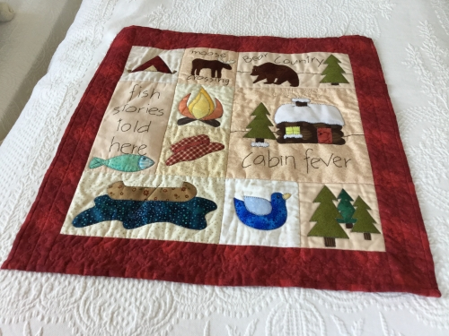 Quilt with burgundy border made of squares depicting aspects of woods: A cabin in show, fir trees, bears, a moose, tent and campfire